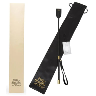 a black riding crop with gold accents and a black wrist strap,, shown with its black storage bag that has the words fifty shades of grey written in gold