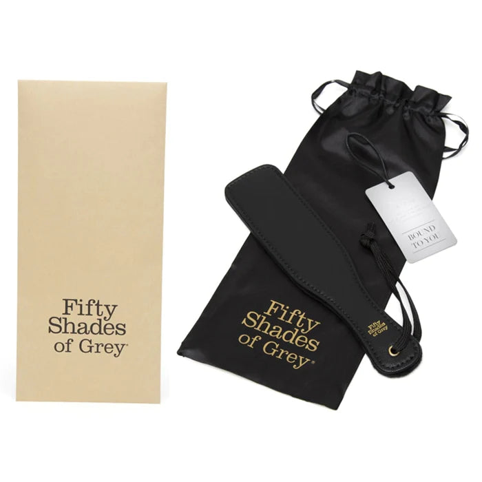 a black paddle with a black wrist strap and a black storage bag. Both have the words fifty shades of grey in gold writing. Shown with its beige packaging.