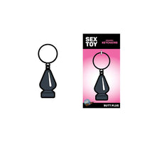The image shows a pink metal butt plug keychain with a ring on top. It is displayed both on its own and in packaging that reads 'SEX TOY' and 'BUTT PLUG'.