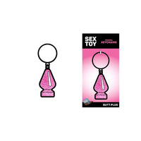 The image shows a pink metal butt plug keychain with a ring on top. It is displayed both on its own and in packaging that reads 'SEX TOY' and 'BUTT PLUG'.