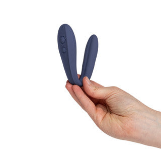 A hand holding a small, dark blue silicone personal massager with two curved prongs against a plain white background.