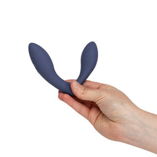 A hand holding a dark blue silicone dual-stimulation vibrator against a plain white background.