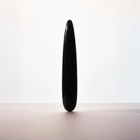 A single black teardrop-shaped stone or ceramic object stands upright on a plain light surface against a white background.