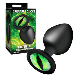 Black silicone butt plug with green eye design next to its packaging on a white background
