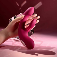 A hand holds a bright pink silicone personal massager with dual curved arms and textured grips against a dark pink and purple studio background with geometric light effects.