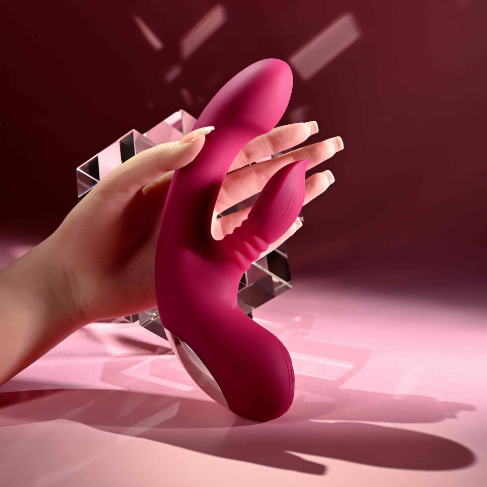 A hand holds a bright pink silicone personal massager with dual curved arms and textured grips against a dark pink and purple studio background with geometric light effects.