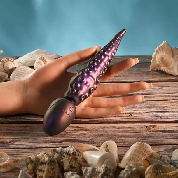 Hand holding a purple and black vibrator with seashells on a wooden surface