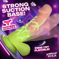 Promotional graphic for a K-pop style suction base dildo, showing a translucent green base with a realistic flesh-toned shaft and glowing green crackle-style lines. Large text reads 'STRONG SUCTION BASE!' and 'HARNESS COMPATIBLE,' with 'FIRM YET FLEXIBLE!' and 'KPOP IDOLS' callouts on a purple neon background.