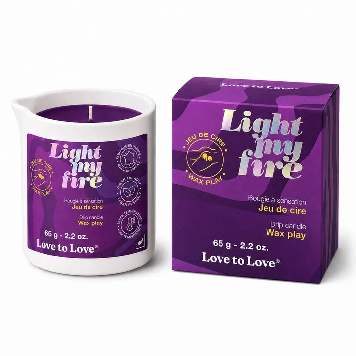 A white wax candle in a white glass vessel with a purple label, shown next to its purple retail box. The packaging and label feature the text 'Light my fire' and 'Love to Love' with additional product details.