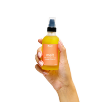Hand holding a bottle of D:C 'melt' massage and bath oil against a white background