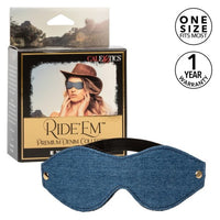A denim blue eye mask with metal studs, displayed in front of its packaging which features an image of a woman wearing a similar eye mask. The packaging also has the Cal Exotics brand logo and mentions the product collection.