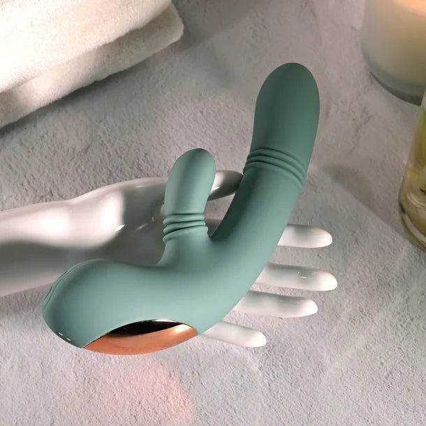 A teal silicone personal massager with a rose gold metallic base, displayed on a white mannequin hand over a textured light gray surface.