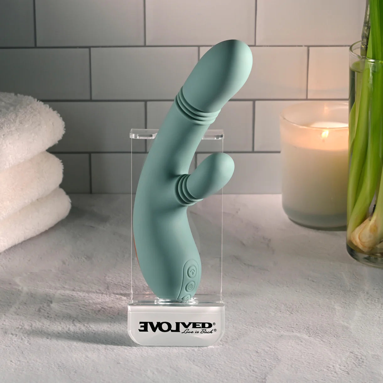 A mint green curved personal massager standing upright in a clear acrylic display case on a countertop. The case base is printed with the brand name 'EVOLVED Love by Revv'.