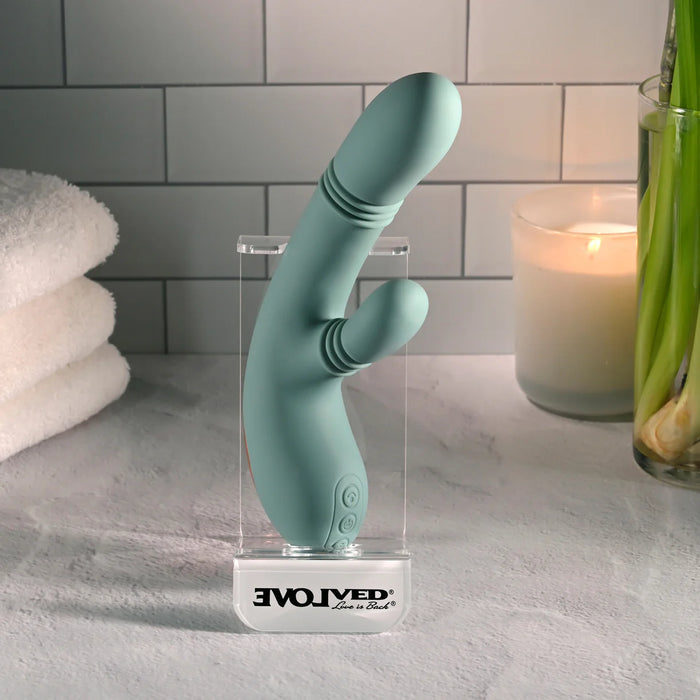 A mint green curved personal massager standing upright in a clear acrylic display case on a countertop. The case base is printed with the brand name 'EVOLVED Love by Revv'.