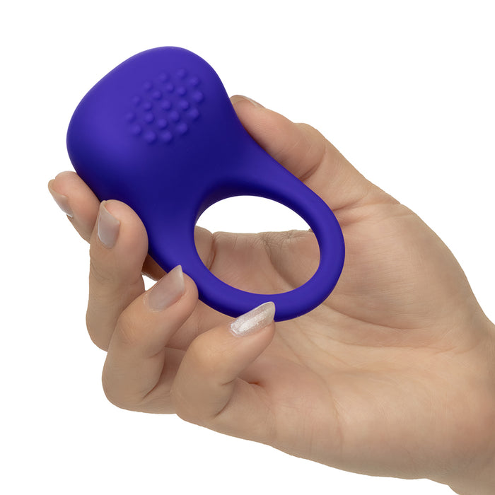 A hand holding a small purple silicone personal massager with a textured, dotted surface and a ring-shaped opening, shown against a plain white background.