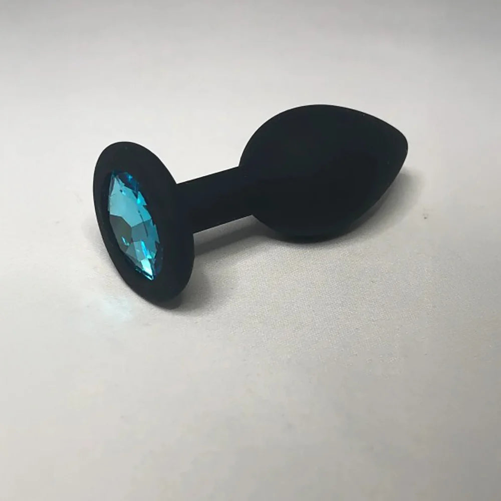 A black silicone butt plug with a faceted light blue gemstone set in a circular base, photographed on a neutral light gray background.