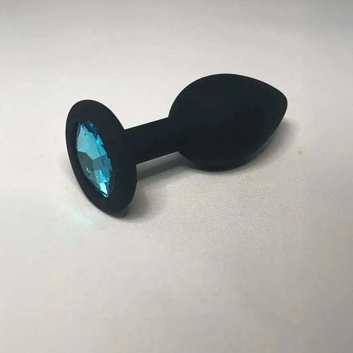 A black silicone butt plug with a faceted light blue gemstone set in a circular base, photographed on a neutral light gray background.