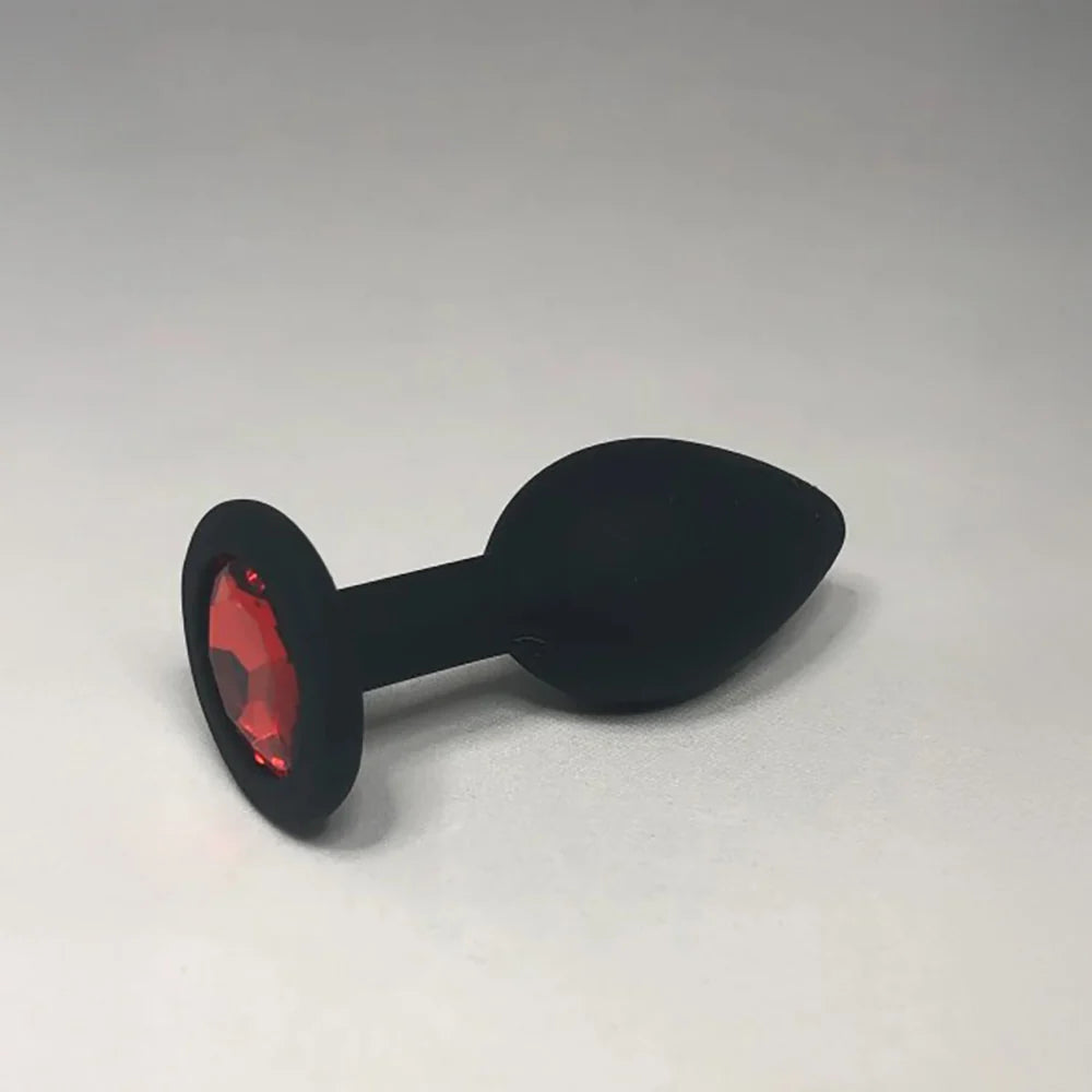 A black silicone butt plug with a faceted red gemstone set in a circular base, photographed on a neutral light gray background.