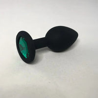 Black silicone butt plug with a faceted green gemstone insert in the base, shown on a light gray surface.