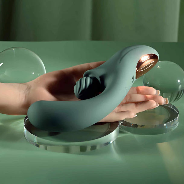 A hand holds a curved, matte sage-green personal massager with a rose-gold metallic interior, resting on clear acrylic stands against a soft green studio background.