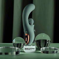 A sleek, curved personal massager in a matte sage green color stands on a clear acrylic display stand. The stand features a white base with the brand name 'EVOLVED' printed in black.