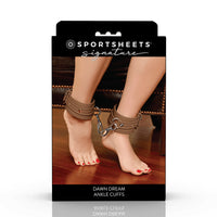 Sportsheets Signature 'Dawn Dream' ankle cuffs in a brown faux-leather material with silver metal hardware, shown on their feet with a chain connecting them, packaged on a black retail card.