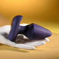 A dark purple silicone anal plug with a curved, textured shaft and a glossy, curved base, displayed on a white hand against a warm yellow gradient background.