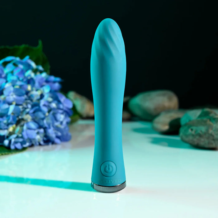 Turquoise vibrator on a light surface with blurred blue flowers and greenery in the background