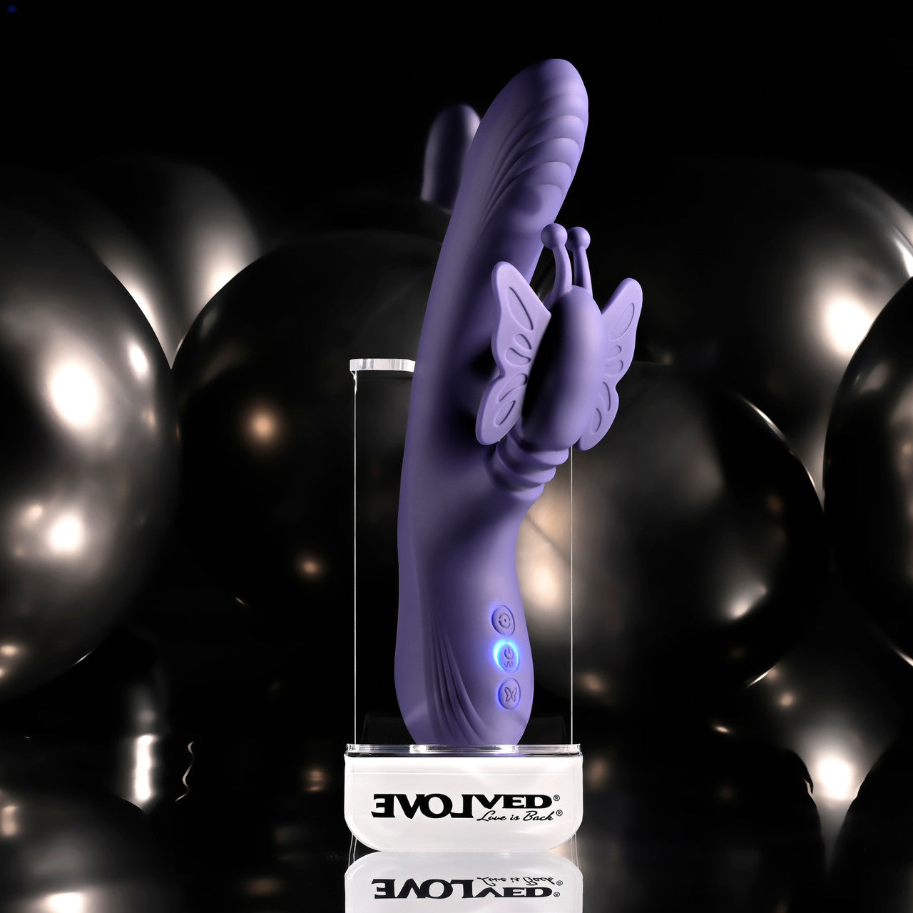 A purple silicone adult novelty massager with a butterfly accent, displayed on a clear stand with a white base printed with the brand name 'EVOLVED Live is Black'. The background consists of dark, reflective metallic balloons against a black backdrop.