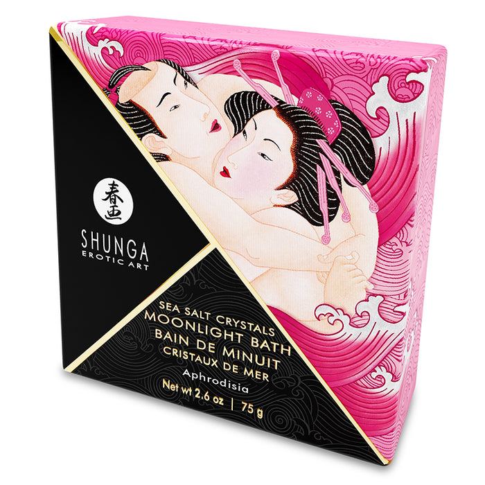 A square container of Shunga Dead Sea salt bath crystals with a graphic of a woman in a pink robe on the label. A rose and scattered bath crystals are shown in the foreground.