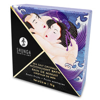 A square container of Shunga Dead Sea salt bath crystals with exotic fruit scent, accompanied by an illustration of a woman and various fruits like grapes and apples. A sea shell scoop is also shown beside the container.