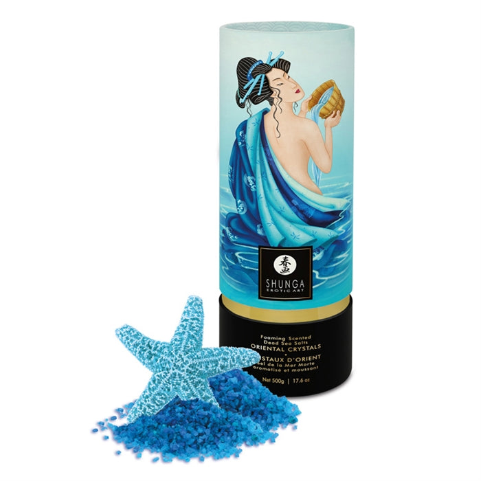 A cylindrical container of Foaming Scented Dead Sea Salt with a graphic of a woman in a bathtub on the label, alongside a blue starfish and a scoop of blue salt crystals.