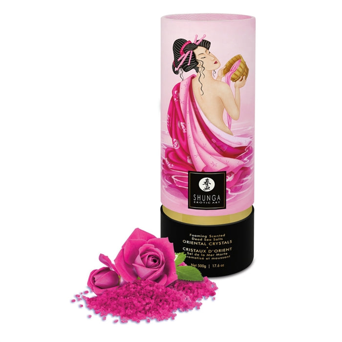 A cylindrical container of Shunga Dead Sea salt bath crystals with a graphic of a woman in a pink robe on the label. A rose and scattered bath crystals are shown in the foreground.