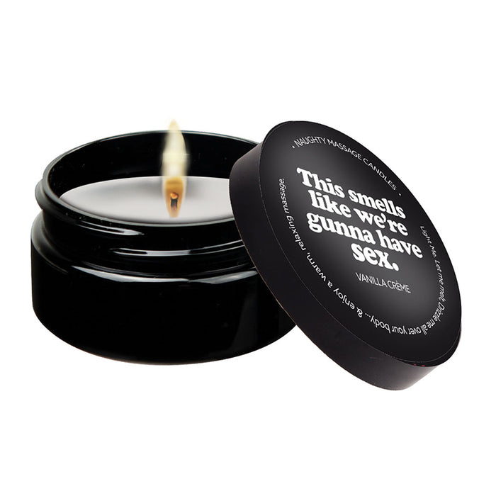 A vanilla massage candle with a label that reads 'This smells like we're gunna have sex', set in a black container. The candle is lit, and there is a pool of melted oil beside it.