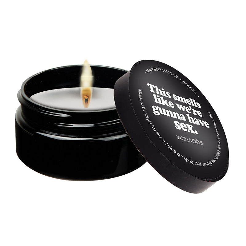 A vanilla massage candle with a label that reads 'This smells like we're gunna have sex', set in a black container. The candle is lit, and there is a pool of melted oil beside it.